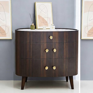Puede incluir: Cómoda de madera marrón oscuro con tres cajones y tiradores dorados en forma de flor. La superficie superior es blanca con un patrón de mármol. La cómoda tiene un diseño curvo y se apoya en cuatro patas cónicas. Objetos decorativos encima.