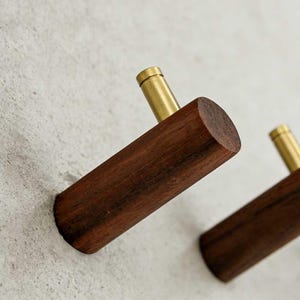 May include: Two wooden wall hooks with brass accents. The hooks are made of dark brown wood and have a cylindrical shape. The brass accents are at the top of each hook. The hooks are mounted on a light gray wall.