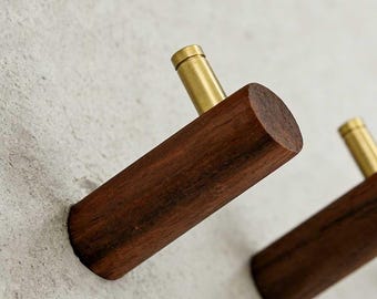 May include: Two wooden wall hooks with brass accents. The hooks are made of dark brown wood and have a cylindrical shape. The brass accents are at the top of each hook. The hooks are mounted on a light gray wall.