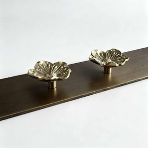 Op de afbeelding: Twee gepolijste, bloemvormige messing knoppen gemonteerd op een rechthoekige, donkerbronskleurige metalen balk. Elke knop heeft een gedetailleerd, meerbladig bloemontwerp met een centrale stamper als functioneel hardware-element.