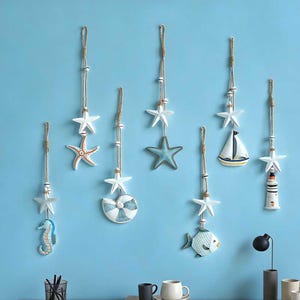 May include: A collection of nautical-themed wall decorations, including starfish, a seahorse, a sailboat, a lighthouse, a seashell, and a fish. Each piece hangs from a rope with wooden beads. The ornaments are in white, blue, and orange, evoking a seaside theme.