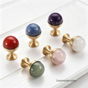 May include: Six spherical cabinet knobs featuring polished stone tops in red, blue, dark purple, light pink, white, and sage green, each set into a brushed brass-toned metal base. The text "Customizable colors" is printed in the bottom right corner.