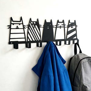 Puede incluir: Perchero de pared de metal negro con cinco siluetas de gatos estilizados. Cada gato tiene un diseño único, con rayas o colores lisos. Una chaqueta azul y una mochila gris cuelgan de los ganchos debajo de los gatos.