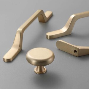 Könnte beinhalten: Eine Sammlung goldfarbener Schrankbeschläge, darunter zwei gebogene Griffe, ein kleinerer Griff und ein runder Knopf. Die Beschläge haben eine matte Oberfläche und sind auf einer grauen Oberfläche angeordnet.