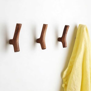 May include: Three wooden wall hooks, crafted to resemble branches, are mounted on a white wall. The hooks are a natural brown colour. A yellow garment hangs from one of the hooks. The hooks are evenly spaced.