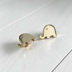 May include: Two gold-toned metal cabinet knobs shaped like stylised elephants. Each knob features a smooth, reflective surface with a small circular indentation representing the eye, designed for attachment to furniture or cabinetry doors.