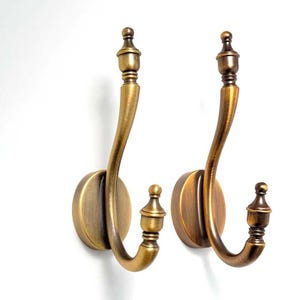 Pode incluir: Dois ganchos de parede com acabamento em latão antigo escovado. Cada gancho apresenta uma placa de base circular, um braço longo curvo e um remate decorativo no topo. O design inclui um gancho secundário menor na base, também rematado com um adorno.
