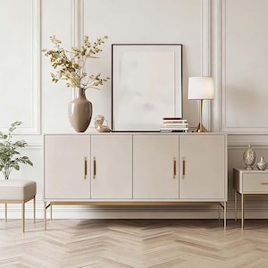 Könnte beinhalten: Ein cremefarbenes modernes Sideboard mit vier Türen und goldfarbenen Beinen. Das Sideboard ist mit einem gerahmten Kunstwerk, einer Lampe, Büchern und dekorativen Gegenständen versehen. Eine Vase mit Zweigen befindet sich links.