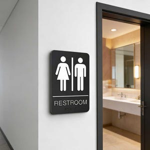 May include: Black restroom sign with white gender symbols and the word "RESTROOM" in white text. The sign also includes Braille text. The sign is mounted on a white wall.