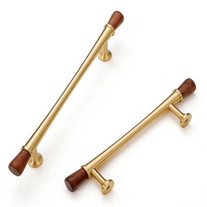 May include: Two brass cabinet pulls with dark wood accents. The pulls have a cylindrical design with a smooth, polished finish. The wood accents are at each end of the pull, adding a touch of warmth to the design. The pulls are designed to be mounted on a cabinet door or drawer.
