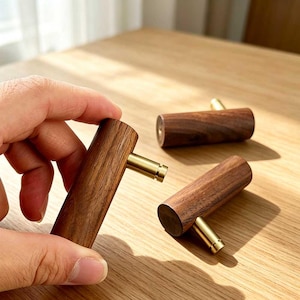 May include: Three cylindrical cabinet knobs made of dark brown wood with a visible grain pattern. Each knob features a polished gold-toned metal post extending from the center of the wooden cylinder, designed for attachment to furniture or doors.