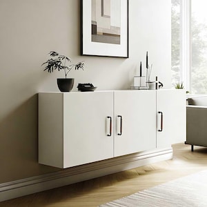 Puede incluir: Un mueble flotante blanco con tres puertas y tiradores marrones. El mueble está montado en una pared beige, con una pequeña planta y objetos decorativos encima. Una obra de arte enmarcada cuelga encima del mueble.
