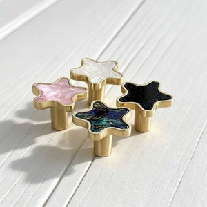 Puede incluir: Cuatro pomos para armarios en forma de estrella con bases de metal dorado. Cada pomo presenta una incrustación única: uno es blanco nacarado, uno es rosa suave, uno es negro profundo con brillo sutil y otro tiene un patrón azul y verde.