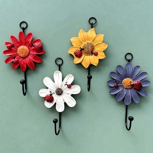 May include: Four wall hooks featuring flower designs in red, yellow, white, and purple. Each flower has a textured centre and small red ladybug accents. A black metal hook extends from the bottom of each flower, and a circular loop is at the top.