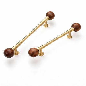 Könnte beinhalten: Zwei Schrankgriffe mit modernem Design. Jeder Griff hat eine dunkelbraune Holzkugel an jedem Ende einer goldfarbenen Stange. Die Griffe haben ein schlichtes, elegantes Design, das sich für Schubladen oder Schränke eignet.