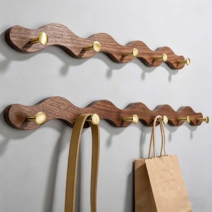Può includere: Due attaccapanni da parete in legno con un design ondulato. Ogni attaccapanni ha più ganci color ottone. Un attaccapanni ha un cinturino in pelle marrone chiaro e una borsa di carta marrone appesa. Il legno è marrone scuro.