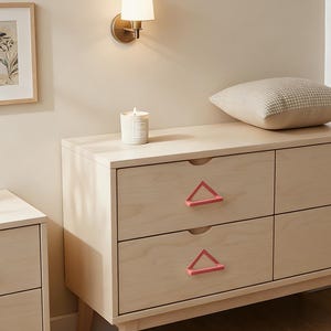 May include: A light-colored wooden dresser with six drawers, featuring pink triangle-shaped drawer pulls. A lit candle sits on top, and a decorative pillow rests on the right. A wall sconce and framed artwork are visible in the background.