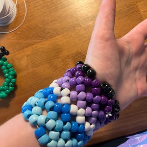 May include: A handmade beaded cuff bracelet in shades of blue, white, and purple, worn on a wrist. The cuff is made of small, round beads. A green beaded wreath and a spool of white thread are also visible.