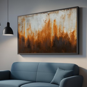 以下が含まれることがあります： 暗いフレームに入った抽象芸術作品。この作品は、白、茶色、オレンジの色合いが混ざり合ったテクスチャのある表面が特徴で、素朴で風化した外観を作り出しています。青いソファの上にある灰色の壁に掛けられています。