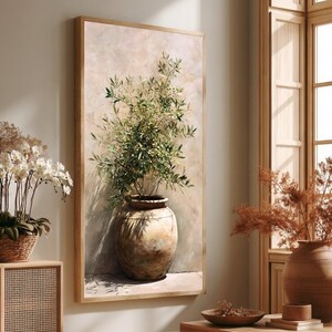 Op de afbeelding: Een ingelijste verticale wandkunstprint met een olijfboom in een rustieke, verweerde aarden pot. Het schilderij bevat zachte, neutrale beigetinten met groen gebladerte en schaduwen tegen een gestructureerde, lichtgekleurde achtergrond.