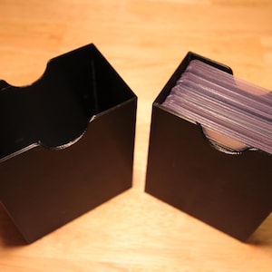 May include: Two black card storage boxes. One box is empty, and the other is filled with clear card sleeves. The boxes have a curved cutout on the front for easy access to the cards. The boxes are sitting on a wooden surface.