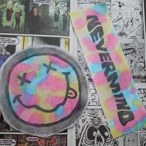 Puede incluir: Dos parches de tela teñidos anudados. Uno es un parche redondo con una cara sonriente con una X por ojos. El otro es un parche rectangular con la palabra "NEVERMIND" escrita en negro.