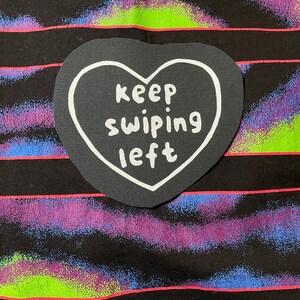 以下が含まれることがあります： ストライプの背景に「keep swiping left」という白い文字が書かれた黒いハート型のワッペン