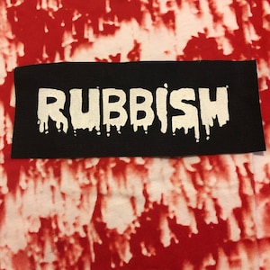 Pode incluir: Um remendo preto com a palavra "RUBBISH" em letras brancas que pingam. O remendo está sobre um fundo tie-dye vermelho e branco.