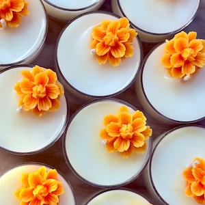 May include: A collection of round glass containers filled with smooth white wax, each topped with a single vibrant orange flower-shaped wax accent. The candles are arranged in a cluster, showcasing the consistent design of the floral wax toppers.