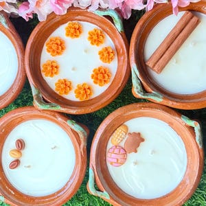 May include: Five round terracotta bowls filled with white wax candles. The candles are decorated with various wax embeds including cinnamon sticks, orange flowers, small brown oval shapes, and miniature colorful baked goods like pan dulce.