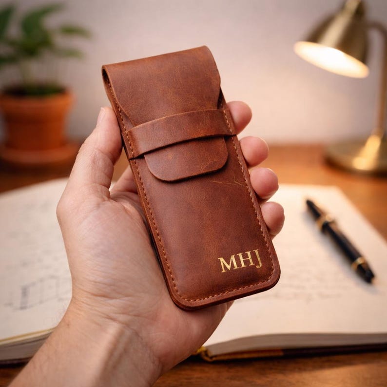 May include: A brown leather pen case with a flap closure and strap. The case has the initials "MHJ" in gold lettering. The case is held in a hand, with a pen and notebook in the background.