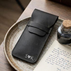 May include: A black leather pen case with a flap closure and a strap. The case is embossed with the initials "S&hearts;K". A small glass ink bottle with a cork stopper and a handwritten letter are next to the case.