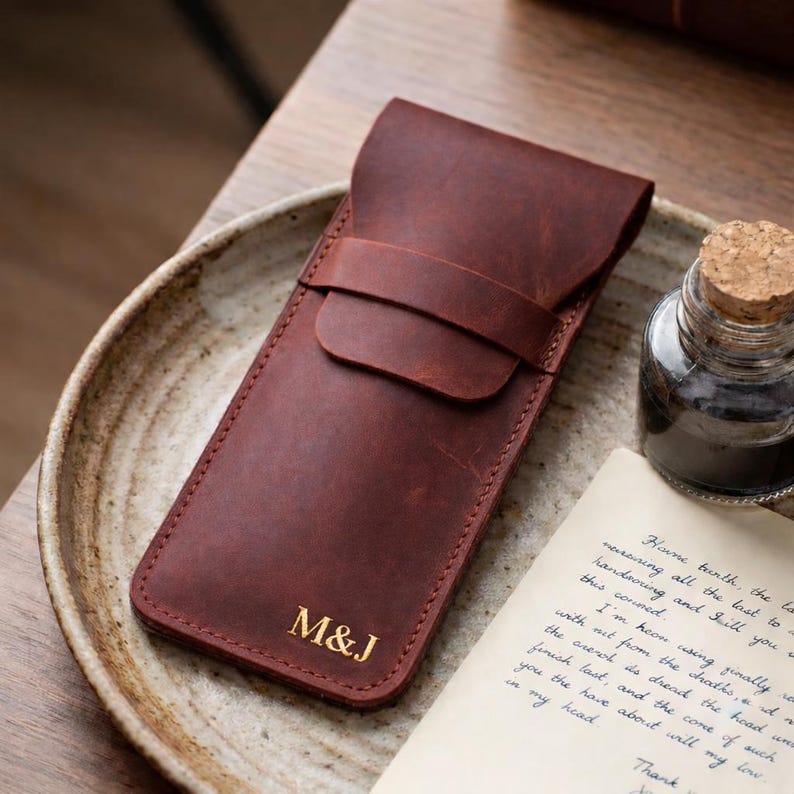 May include: A brown leather pen case with a flap closure and a strap. The initials "M&J" are embossed in gold on the front. The case is on a ceramic plate next to a glass ink bottle and a handwritten letter.