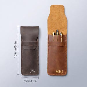 May include: Two leather pen cases, one closed and one open, displaying pens. The closed case is dark brown with the initials "JW". The open case is tan, with three gold pens visible. The cases are 6.5 inches tall and 2.7 inches wide.