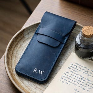 May include: A navy blue leather pen case with a flap closure and a strap. The initials "R.W." are embossed in white on the front. A bottle of ink and a handwritten letter are next to the case.