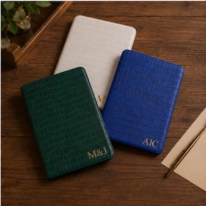 Puede incluir: Tres cuadernos rectangulares con cubiertas texturizadas en crema, verde oscuro y azul real. El cuaderno verde está grabado con M&J, el azul con AIC y el crema con M. Cada cubierta presenta un patrón de cuadrícula y letras doradas.