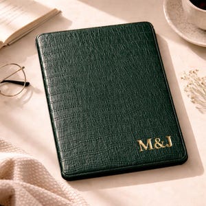 May include: A dark green journal with a crocodile-effect texture. Gold initials "M&J" are embossed on the lower right corner. The journal is rectangular and lies on a light surface, with a book and glasses visible in the background.