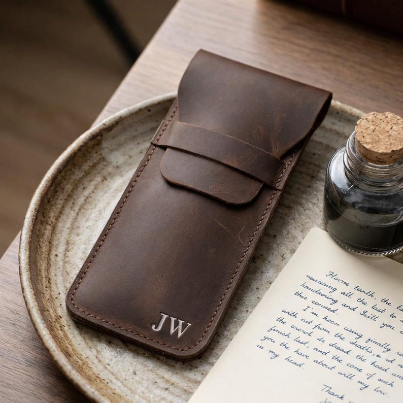 May include: A brown leather pen case with a flap closure and a strap. The initials "JW" are embossed on the front. The case is on a ceramic plate, next to an ink bottle and a handwritten letter.