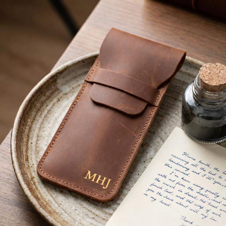 May include: A brown leather pen case with the initials "MHJ" in gold lettering. The case has a flap closure and a stitched border. It is displayed on a ceramic plate with a bottle of ink and a handwritten letter.