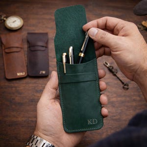 May include: A dark green leather pen case with three pens inside. The case is open, revealing a black and gold pen, and two gold pens. The initials "KD" are embossed on the case. Two other leather pen cases are in the background.