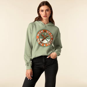 Puede incluir: Sudadera con capucha verde salvia con un gráfico circular que dice "MOM OF THE YEAR". El gráfico presenta hachas cruzadas, montañas y una brújula. La sudadera tiene una capucha con cordón y un bolsillo delantero.