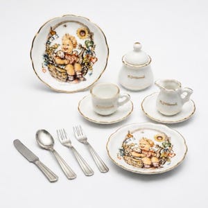 Puede incluir: Un juego de té de porcelana en miniatura que incluye dos platos con ilustraciones de un niño en una cesta, un azucarero con tapa, una jarra de leche y una taza sobre un platillo. El set contiene un cuchillo, una cuchara y dos tenedores de metal plateado, con bordes dorados y la firma M.J. Hummel.