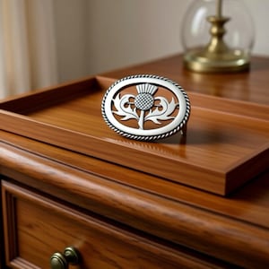 May include: An oval, silver-colored belt buckle featuring a thistle design. The buckle is set against a wooden tray on a wooden dresser. The buckle has a rope-like border and is likely a decorative accessory.