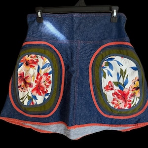 Può includere: Gonna di jeans blu con due grandi toppe floreali ovali. Le toppe presentano fiori rossi, bianchi e blu con bordi verdi e arancioni. La gonna è appesa a una gruccia nera.