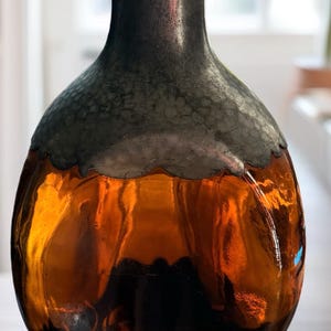 May include: An amber glass bottle with a metal rim and a cork stopper. The bottle has a rounded body and a narrow neck. The metal rim has a scalloped edge. The bottle is likely used for storing liquids.