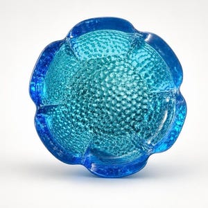 May include: A vibrant blue glass bowl shaped like a flower with five rounded petals. The interior surface features a raised, textured honeycomb pattern that catches the light, while the outer edges are smooth and translucent with a deep blue hue.