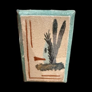 Op de afbeelding: Een rechthoekige keramische tegel met een lichtbeige achtergrond en een turquoise rand. De tegel toont een gestileerde illustratie van een plant met donkergrijze en zwarte bladeren, een kleine witte structuur en bruine accenten.