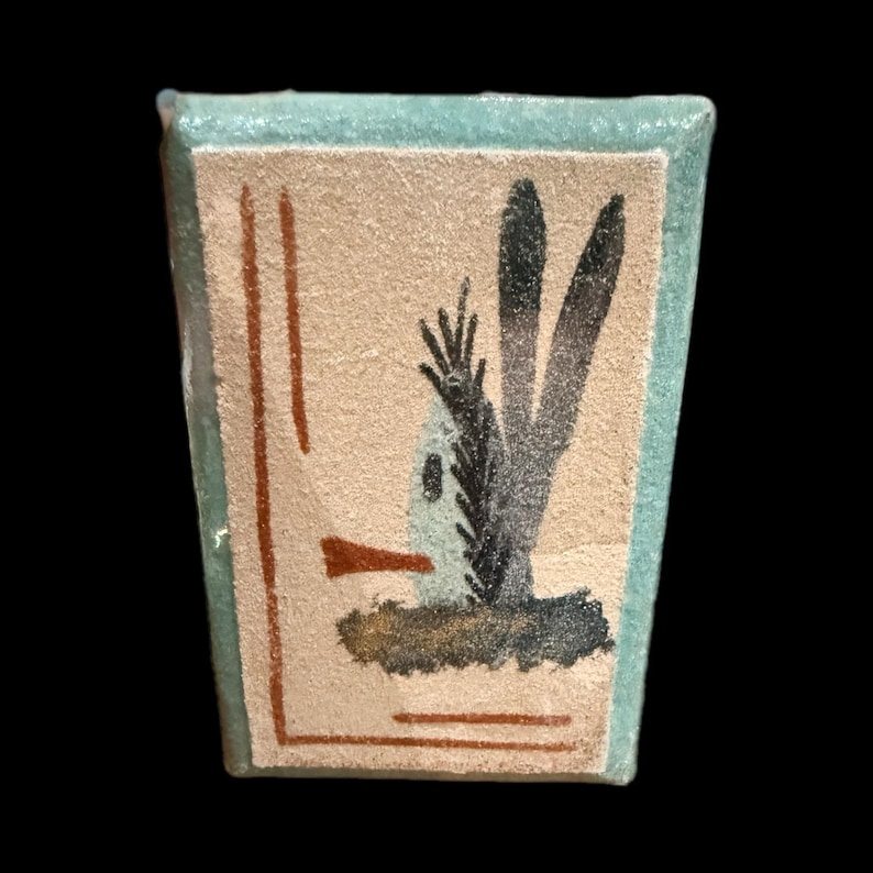 Op de afbeelding: Een rechthoekige keramische tegel met een lichtbeige achtergrond en een turquoise rand. De tegel toont een gestileerde illustratie van een plant met donkergrijze en zwarte bladeren, een kleine witte structuur en bruine accenten.