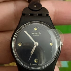 May include: Black Swatch Swiss watch with a dark face, gold accents, and a black strap. The watch face has the Swatch Swiss logo and the text "SWATCH AG 2000".