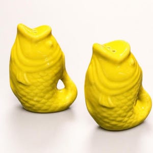 May include: A pair of bright yellow ceramic salt and pepper shakers shaped like fish. Each shaker features a textured scale pattern, a curved tail, and an open mouth design with small holes on the top for dispensing seasoning.